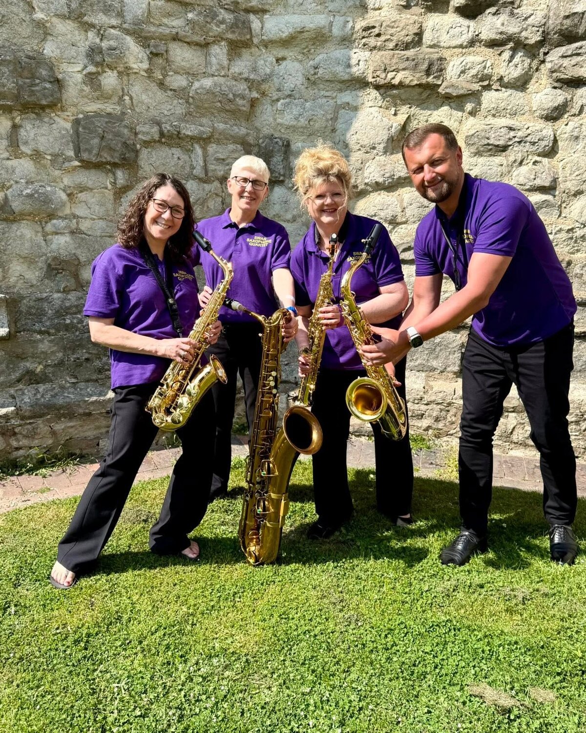 Swan Saxophone Quartet smiling together with their saxophones beside a stone wall